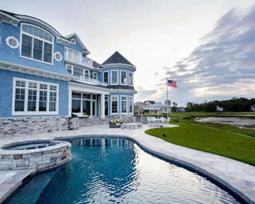 Large blue house with many windows overlooking a backyard pool and hot tub, perfect for outdoor living. An American flag flies near the grassy area and marshland—ideal for enjoying inground pools with scenic views.
