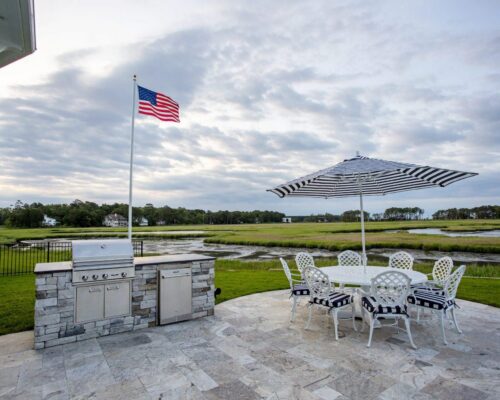 Outdoor patio with a stone grill, round table, striped umbrella, and American flag overlooking marshland under a cloudy sky—perfect for gatherings near outdoor kitchens or luxury pools.