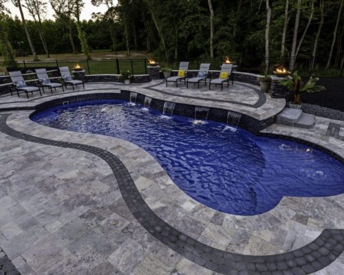Backyard swimming pool with decorative stone deck, water features, lounge chairs, and built-in fire bowls—perfect for relaxing by inground pools or custom spas—surrounded by trees and fencing.