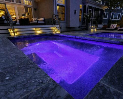 A hot tub with purple LED lights, part of a custom spa, is built into a stone patio in a backyard at dusk, with illuminated steps and the house visible in the background.
