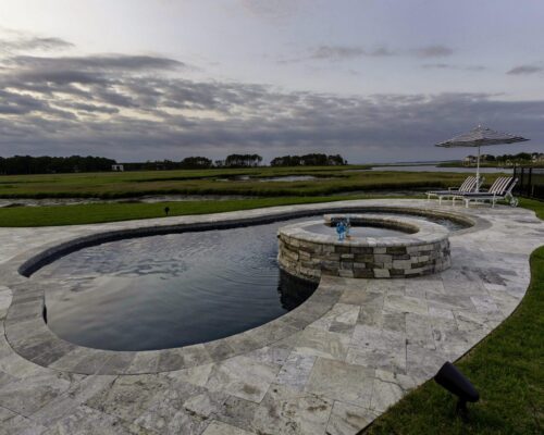 Outdoor living at its best: inground pool with an attached circular hot tub, surrounded by stone decking and overlooking grassy wetlands under a cloudy sky; lounge chairs and umbrella complete this serene custom spa retreat.