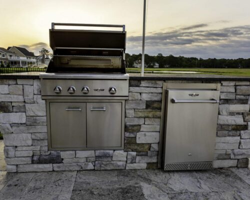 Stainless steel outdoor grill with open lid and a built-in refrigerator, set in a stone island on a patio overlooking fiberglass pools and a grassy landscape at sunset.