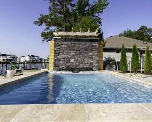 Rectangular inground pool with a stone waterfall feature at one end, surrounded by trees and houses under a clear blue sky—a true oasis for those seeking luxury pools.