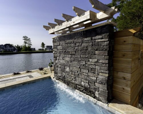 A stone wall water feature with a wooden pergola spills water into a small pool, creating a serene outdoor living space beside a body of water and surrounded by lush landscaping.
