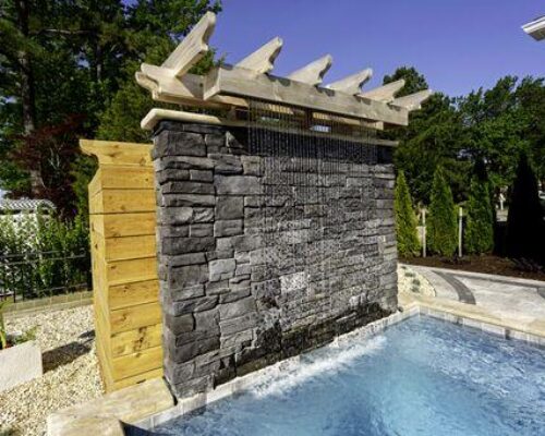 A stone water wall feature with a pergola pours water into a small luxury pool, surrounded by landscaping and trees—perfect for pairing with outdoor kitchens or custom spas.