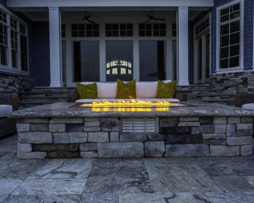 Outdoor patio with a stone fire pit, cushioned sofa with green pillows, wicker chairs, and views of luxury pools in front of a house with large windows and steps.