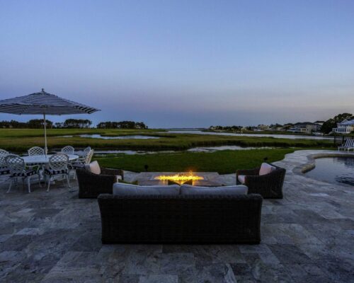 Patio with wicker sofa, chairs, fire pit, and luxury pool beside grassy wetlands at sunset; striped umbrella and dining set to the left.