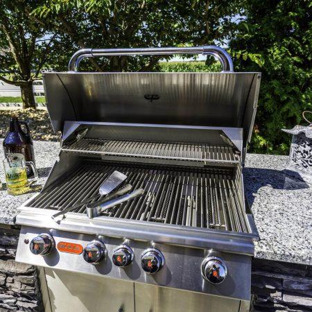 A stainless steel outdoor grill with its lid open, grilling tools on the grates, two mugs with drinks, a bottle, and a lantern on a stone countertop in a sunny backyard—perfect for outdoor kitchens or gatherings by luxury pools.