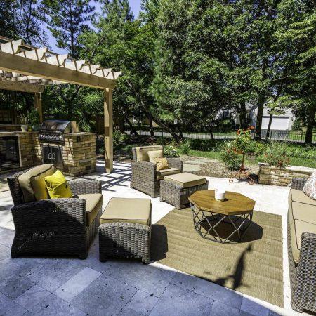Outdoor patio with wicker furniture, beige cushions, a round coffee table, pergola with built-in grill and outdoor kitchen, plus lush greenery in the background on a sunny day.