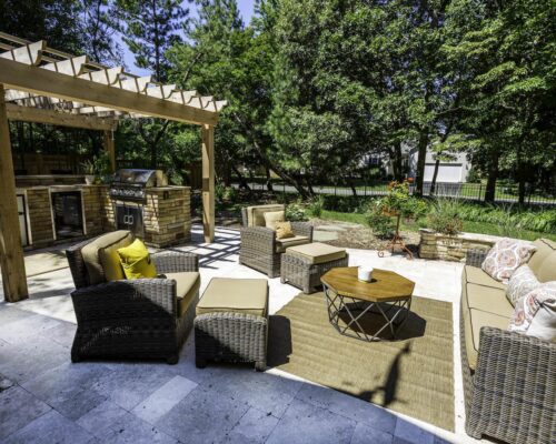 Outdoor patio with wicker furniture, beige cushions, a round wooden table, and a pergola over a built-in grill and custom outdoor kitchen area, surrounded by trees and greenery.
