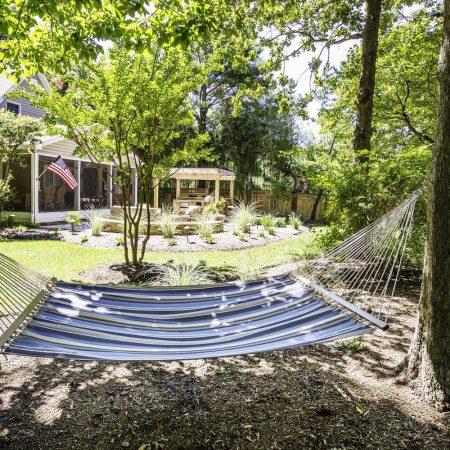 A blue hammock is suspended between two trees in a sunny backyard featuring a house, garden, gazebo, an American flag, and views of luxury pools in the background.