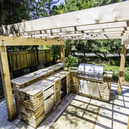 Outdoor kitchen under a wooden pergola with a stone countertop, built-in grill, sink, mini fridge, and surrounding greenery—perfect for enjoying luxury pools or complementing custom spas in your backyard oasis.