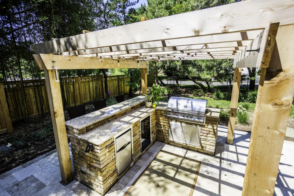 Outdoor kitchen with stone counters, stainless steel appliances, and a grill under a wooden pergola—perfectly paired with luxury pools—surrounded by greenery and a wooden fence.