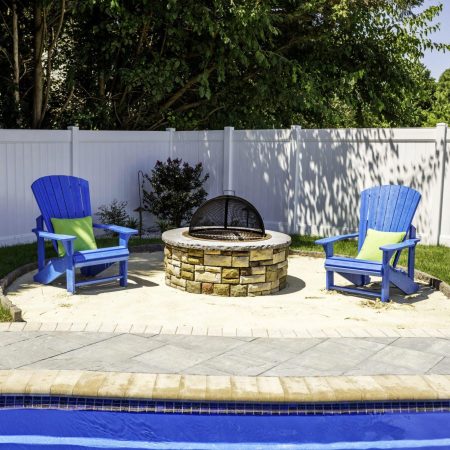 Two blue Adirondack chairs with green pillows sit by a stone fire pit on a patio next to a sparkling fiberglass pool, creating the perfect outdoor living space, with a white fence and trees in the background.
