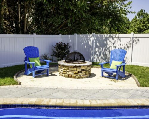 Two blue Adirondack chairs with green pillows sit by a stone fire pit on a patio next to a sparkling fiberglass pool, creating the perfect outdoor living space, with a white fence and trees in the background.