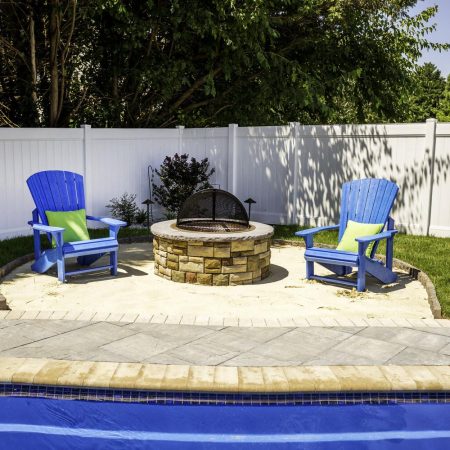 Two blue Adirondack chairs with green pillows sit around a stone fire pit in a fenced backyard beside an inground pool, with trees in the background.