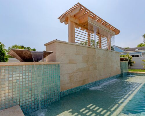 Modern luxury pool with a tiled water feature, cascading waterfall, and wooden pergola overhead—perfect for elevated outdoor living. Surrounded by greenery and residential buildings, it’s an inviting retreat for relaxing outdoors.