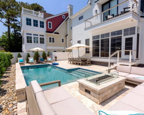 Modern backyard with a rectangular inground pool, lounge area with firepit, dining table with umbrellas, and white multi-story houses in the background—perfect for relaxing or entertaining near stunning fiberglass pools and stylish outdoor kitchens.