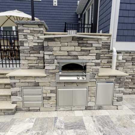 Outdoor kitchen with built-in stainless steel grill and storage set in a stone countertop, next to stone steps and a patio area with an umbrella—perfect for entertaining by inground pools or custom spas.