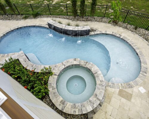 Aerial view of a backyard luxury pool with a built-in custom spa, water features, stone tile decking, and surrounding landscaping.