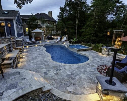 Luxury backyard with a fiberglass pool, custom spa, hot tub, lounge chairs, gazebo, outdoor kitchen and bar, plus a seating area surrounded by trees and upscale homes.