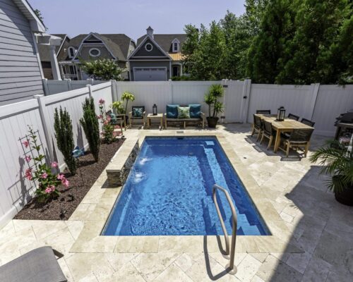 Small rectangular inground pool surrounded by stone tiles, patio furniture, dining table, lush plants, and a white privacy fence on a sunny day—perfect for relaxing or entertaining by your poolside custom spa.