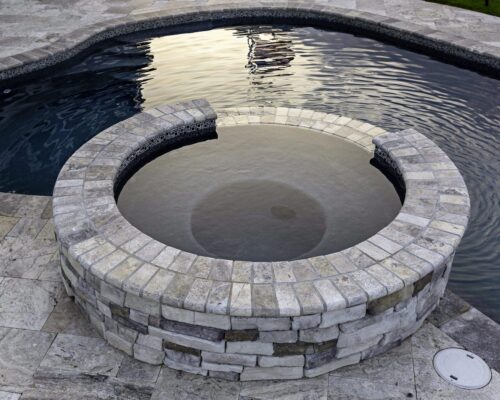 A circular hot tub with stone veneer overflows into an adjacent inground pool, both surrounded by beige stone tile decking.