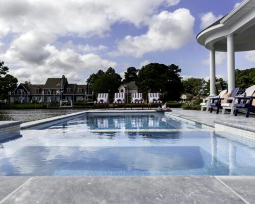 Swimming pool with lounge chairs and Adirondack chairs on a patio, overlooking a lake with houses and trees in the background under a partly cloudy sky—perfect for enjoying outdoor living.