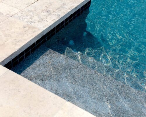 Close-up view of a swimming pool corner with clear blue water, submerged steps, and light-colored stone tiles, perfect for enhancing your outdoor living space with stylish inground pools.