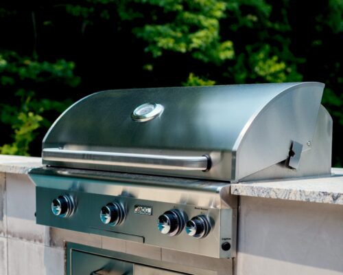 A stainless steel outdoor gas grill with three control knobs and a built-in thermometer, installed in a stone countertop beside luxury pools, with trees visible in the background.