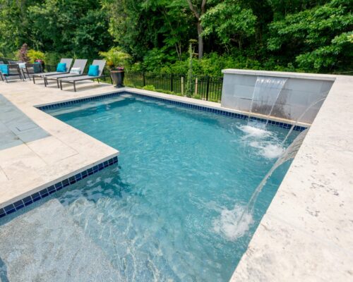 Rectangular backyard swimming pool with a stone deck, water feature, and lounge chairs set beside a backdrop of green trees—perfect for those who appreciate luxury pools and custom spas in their outdoor oasis.