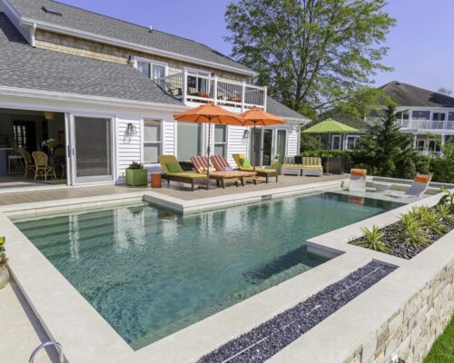 Modern backyard with a rectangular inground pool, poolside lounge chairs under orange umbrellas, and a patio area attached to a two-story house—perfect for those seeking luxury pools and relaxing custom spas.