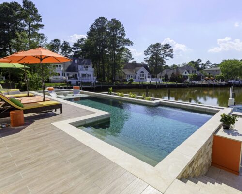 Modern backyard with an inground infinity pool, lounge chairs under an orange umbrella, and a view of waterfront houses and trees along a canal—perfect for outdoor living.
