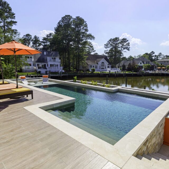 Modern backyard with an inground infinity pool, lounge chairs under an orange umbrella, and a view of waterfront houses and trees along a canal—perfect for outdoor living.