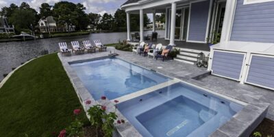 Rectangular inground pool and hot tub beside a blue house with lounge chairs, patio furniture, and a view of a lakeside neighborhood—perfect for relaxing by your fiberglass pool or entertaining near custom outdoor kitchens.