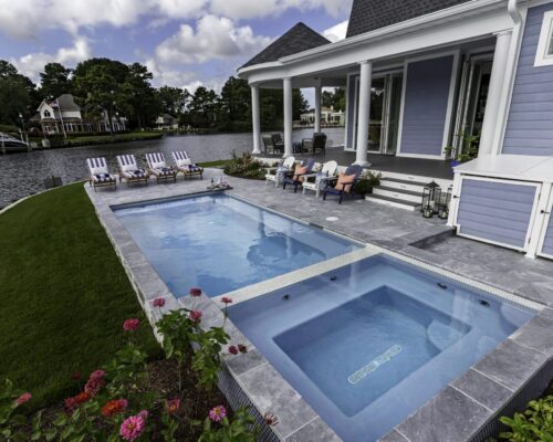 Rectangular inground pool and hot tub beside a blue house with lounge chairs, patio furniture, and a view of a lakeside neighborhood—perfect for relaxing by your fiberglass pool or entertaining near custom outdoor kitchens.