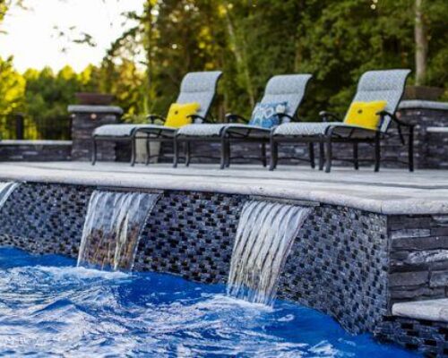 Three lounge chairs with yellow pillows sit on a stone pool deck, overlooking blue water with three small waterfalls. This serene retreat, surrounded by greenery, is the perfect spot to enjoy luxury pools or custom spas in style.