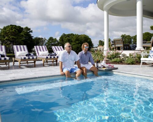 Two men sit on the edge of a swimming pool with their feet in the water. Empty lounge chairs are arranged nearby, and a large house and custom spa are visible in the background.