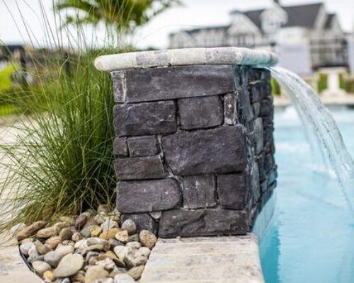 A stone water feature with a waterfall flows into a luxury pool, with ornamental grass and pebbles nearby, creating a serene ambiance for relaxing near custom spas or outdoor kitchens.