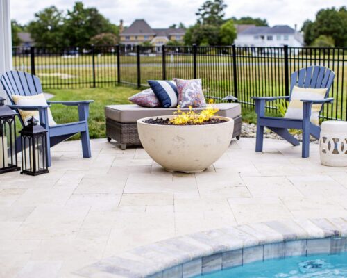 Three blue patio chairs surround a round fire pit beside a sparkling fiberglass pool, with lanterns, cushions, and a grassy yard in the background—perfect for relaxed outdoor living.