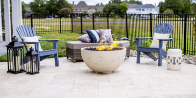 Two blue chairs, a cushioned bench with pillows, and a round fire pit create an inviting outdoor living space on a stone patio near fiberglass pools; two black lanterns sit on the ground.