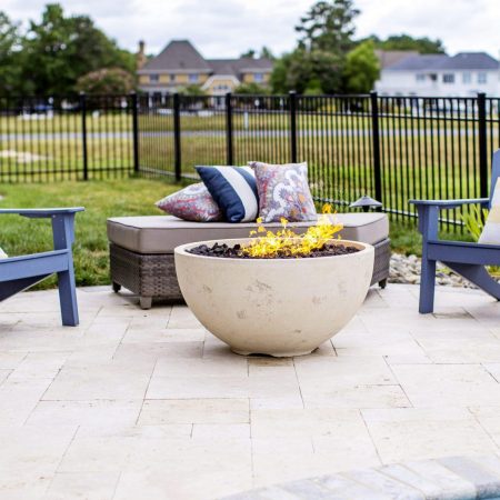 Two blue chairs, a cushioned bench with pillows, and a round fire pit create an inviting outdoor living space on a stone patio near fiberglass pools; two black lanterns sit on the ground.