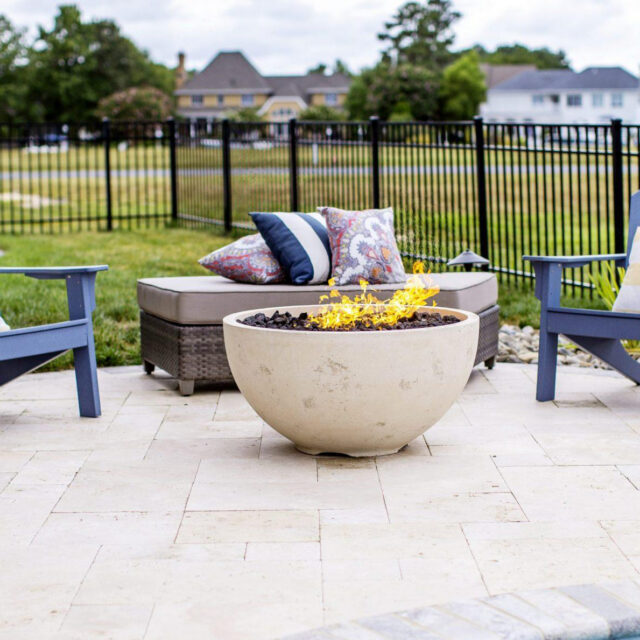 Two blue chairs, a cushioned bench with pillows, and a round fire pit create an inviting outdoor living space on a stone patio near fiberglass pools; two black lanterns sit on the ground.