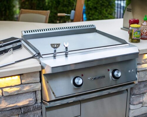 A stainless steel outdoor griddle with grilling tools on top, jars of pickles and sriracha sauce nearby, built into a stone kitchen counter—perfect for enhancing your outdoor living space.