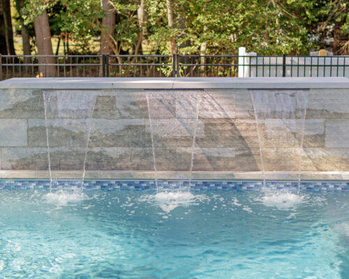 A luxury pool with four streams of water flowing from a stone wall feature into the pool; trees and fencing are visible in the background, perfect for enhancing your outdoor living experience.