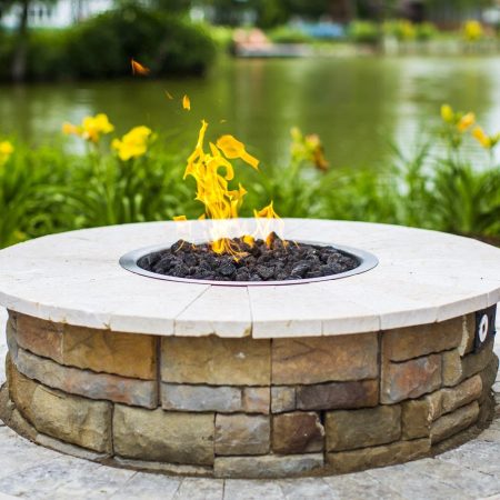 A round stone fire pit with a flame in the center is set on a tiled patio near a pond, surrounded by green plants and yellow flowers—an inviting retreat perfect for relaxing by luxury pools or custom spas.