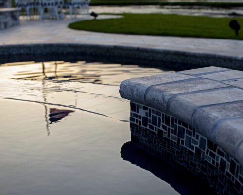 Close-up of the edge of a tiled inground pool at sunset, with water reflecting light and a lawn with patio furniture visible in the background.