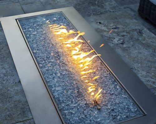 A rectangular outdoor fire pit with flames rising from clear glass rocks, set in a stone patio—perfect for relaxing by your custom spa or enhancing stunning outdoor kitchens.