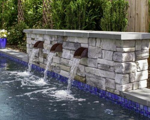 A stone wall with three spouts pours water into a luxury pool lined with blue tiles, surrounded by plants, a wooden fence, and perfect for enjoying custom spas or outdoor kitchens.
