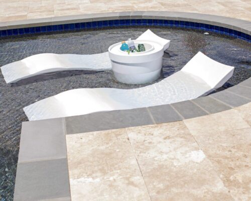 Two white lounge chairs are partially submerged in a shallow inground pool beside a round table holding drinks and sunglasses, with tile flooring surrounding this modern luxury space.